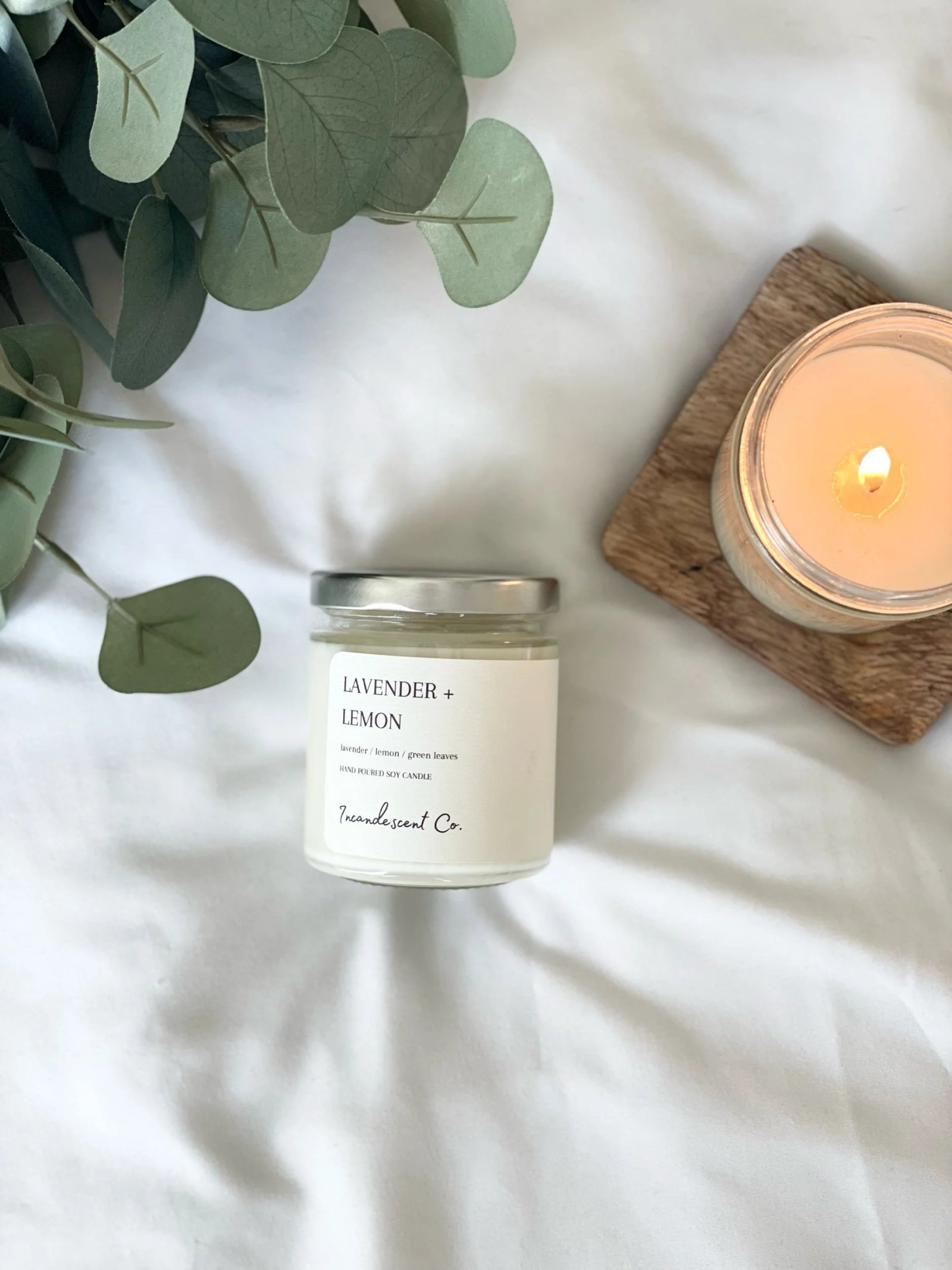 Lavender and lemon scented candle on white background next to a lit candle on a wooden coaster and eucalyptus leaves.