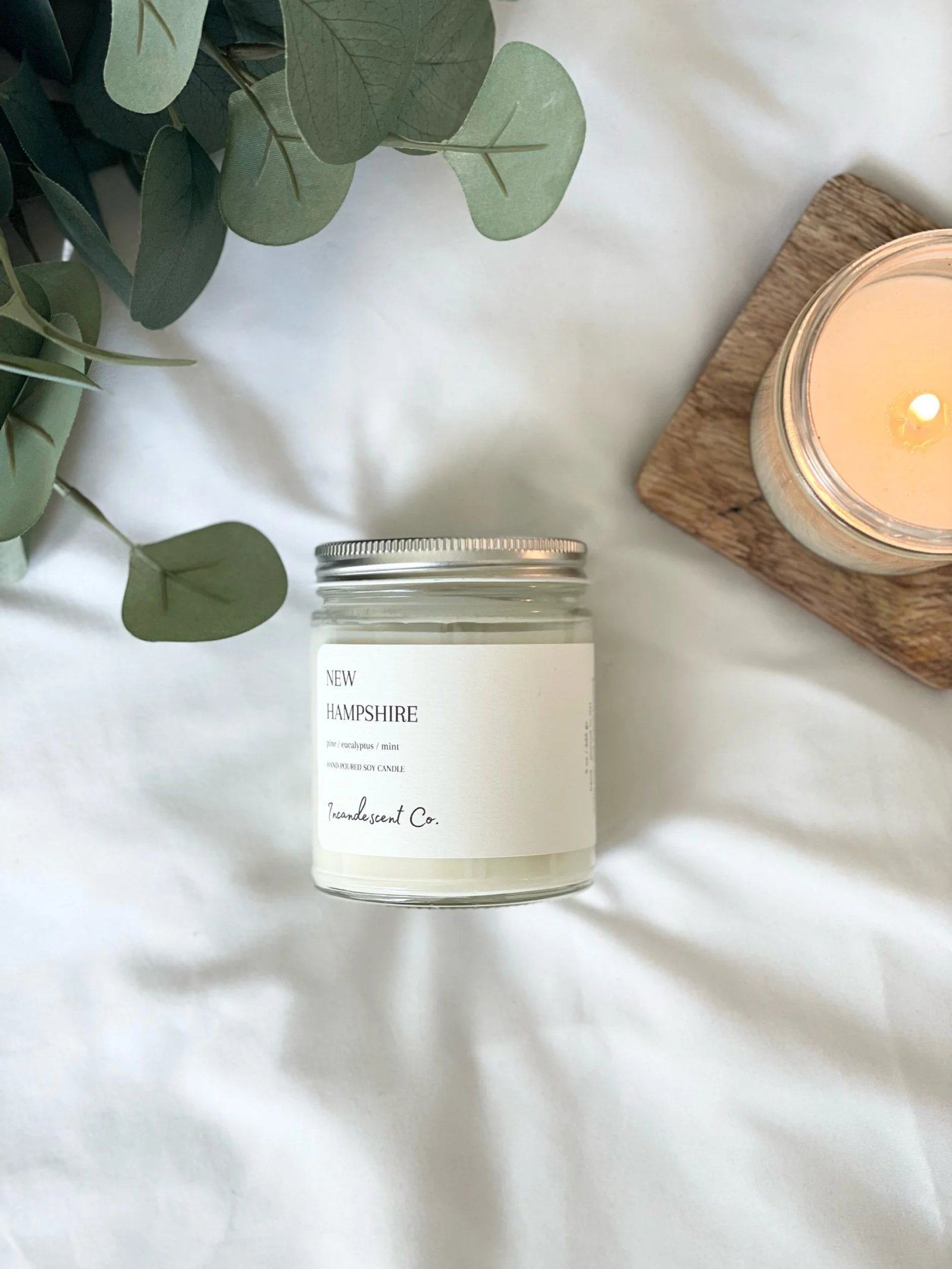 New Hampshire scented candle in a glass jar with eucalyptus and mint, lit candle on wooden coaster, white background.