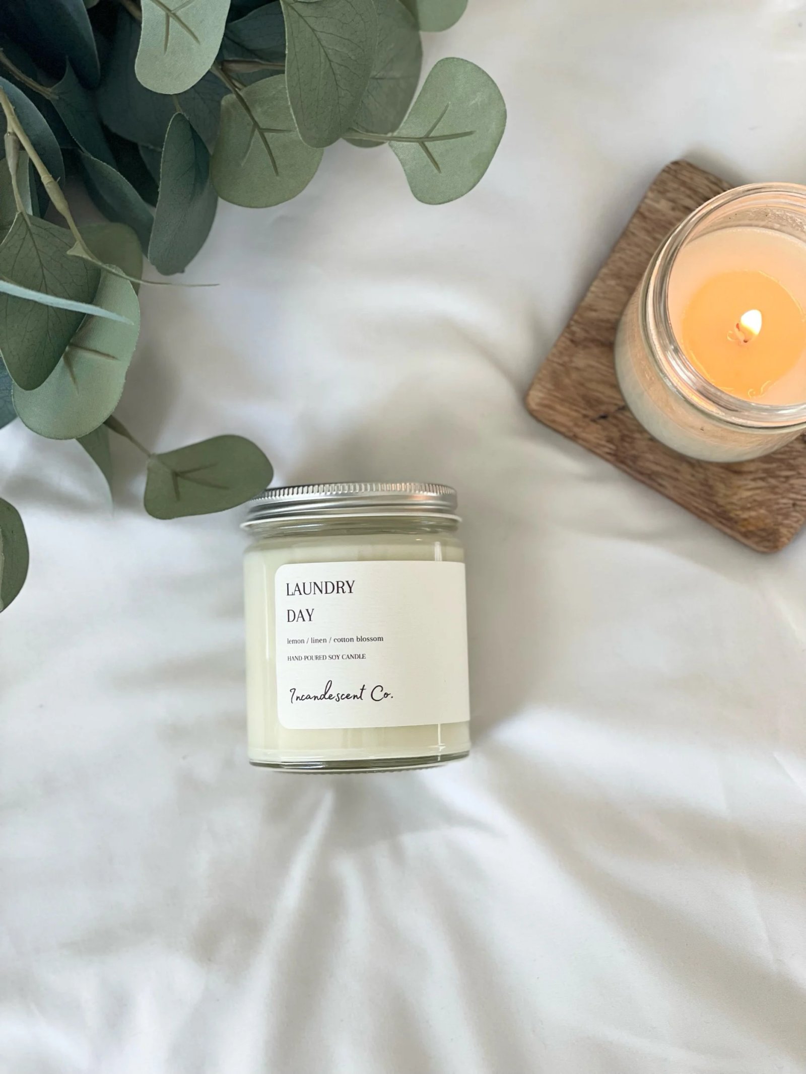 Overhead view of a white soy candle labeled "Laundry Day" in a glass jar on a white surface, next to a lit candle on a wooden coaster. Green leaves are visible in the top left corner.